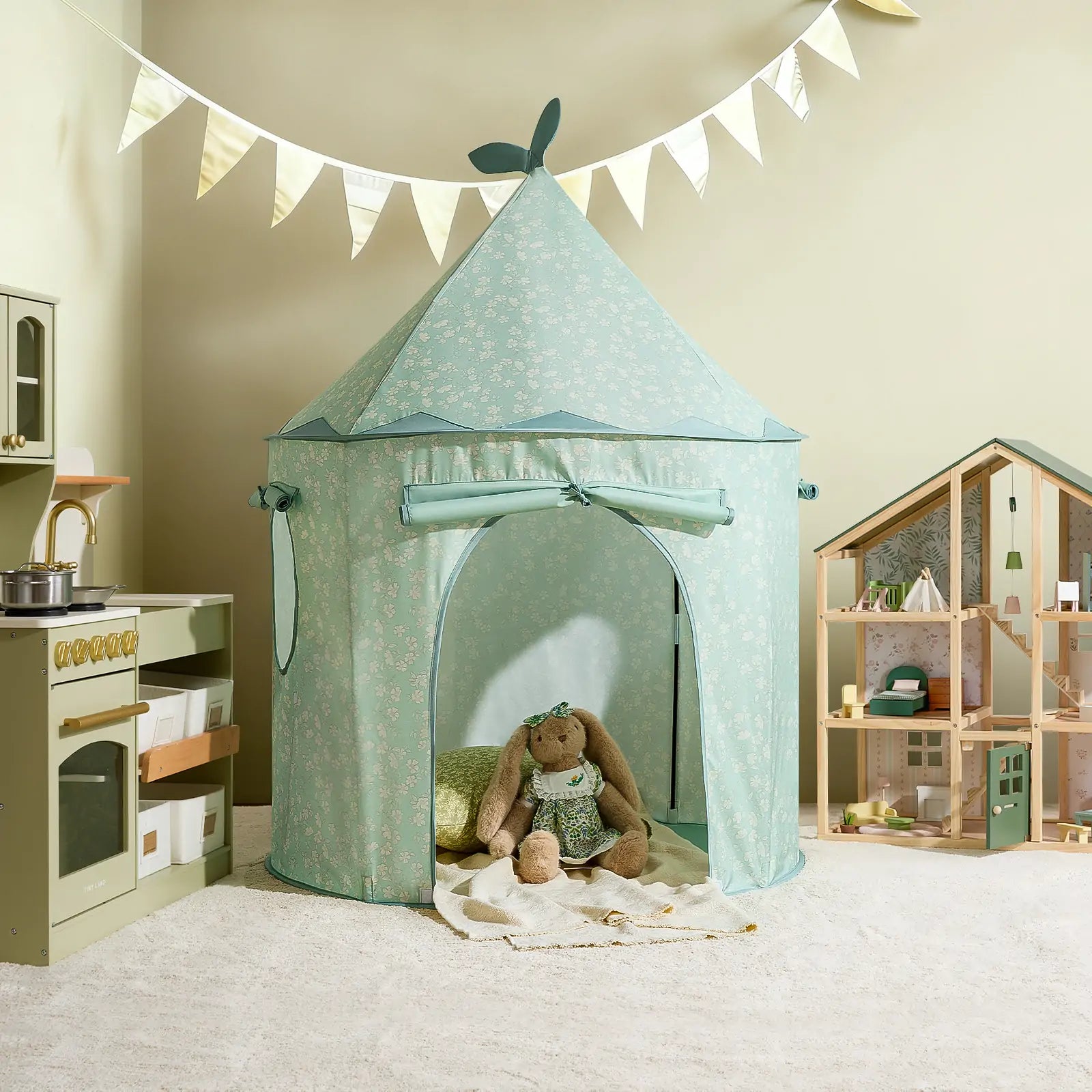 A children’s playroom featuring the Tiny Land® Tiny Sprout Castle Tent in mint-green with a plush bunny inside, a toy kitchen to the left, a dollhouse to the right, and a bunting banner on a cream-colored wall.