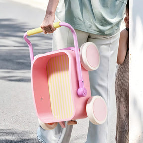 Someone carries the Tiny Land® 3-in-1 Wooden Baby Walker Wagon - Pink by its handle outdoors, while another child stands nearby against a wall.
