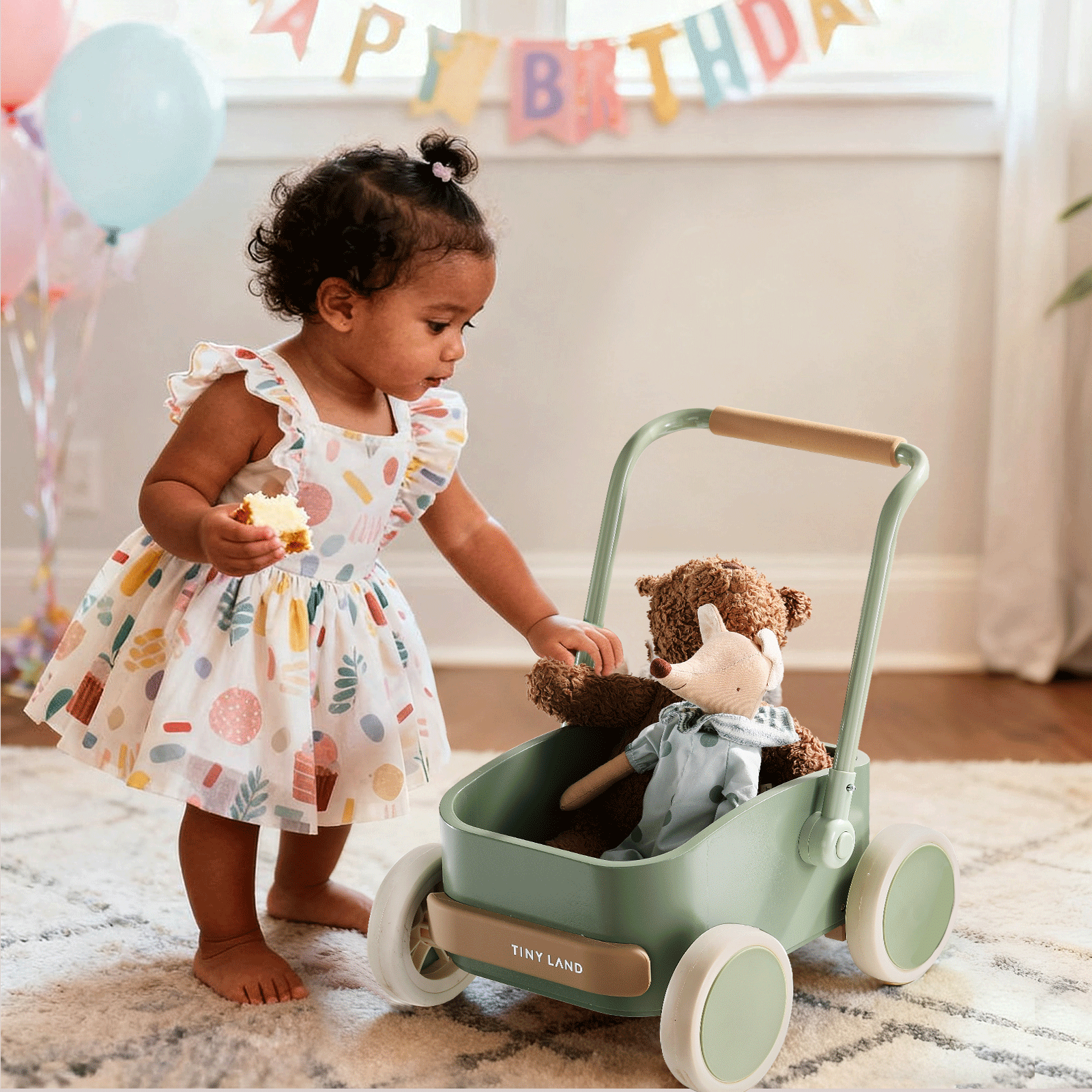 A toddler in a colorful dress holds a cupcake and pushes the Test Tiny Land® JoyRide Baby Walker with Blocks - Sage, which has a teddy bear inside. Birthday decorations and balloons are in the background.