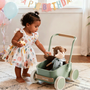 A toddler in a colorful dress holds a cupcake and pushes the Test Tiny Land® JoyRide Baby Walker with Blocks - Sage, which has a teddy bear inside. Birthday decorations and balloons are in the background.
