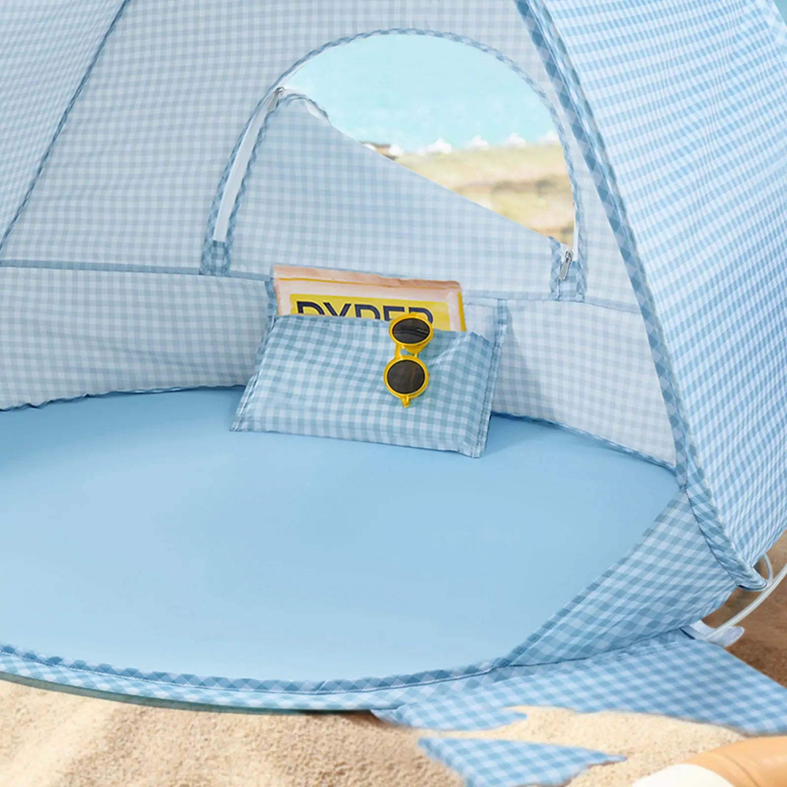 The Tiny Land® One-Stop Beach Toy & Tent Set in light blue gingham sits on the sand, complete with a matching pillow, yellow sunglasses, and magazine. Relax under this UV-protective tent and enjoy your sea view in comfort.