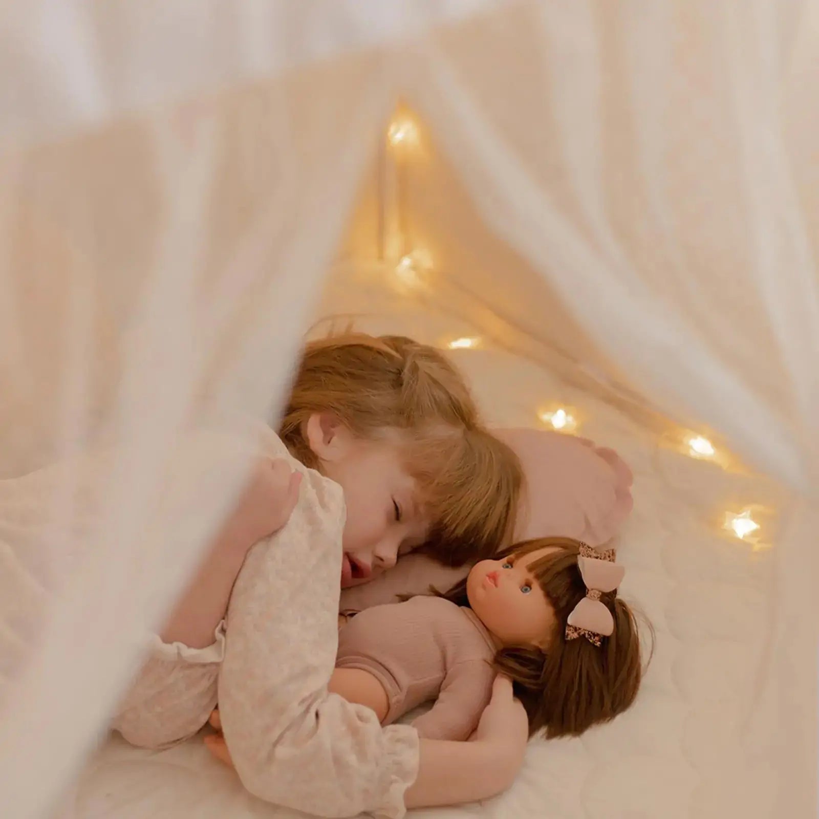A child sleeping inside a Tiny Land® Cozy Nook – GentleSnug Chair & Playhouse Tent, holding a doll and illuminated by string lights.|White