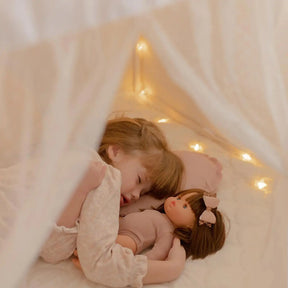 A child sleeping inside a Tiny Land® Cozy Nook – GentleSnug Chair & Playhouse Tent, holding a doll and illuminated by string lights.|White