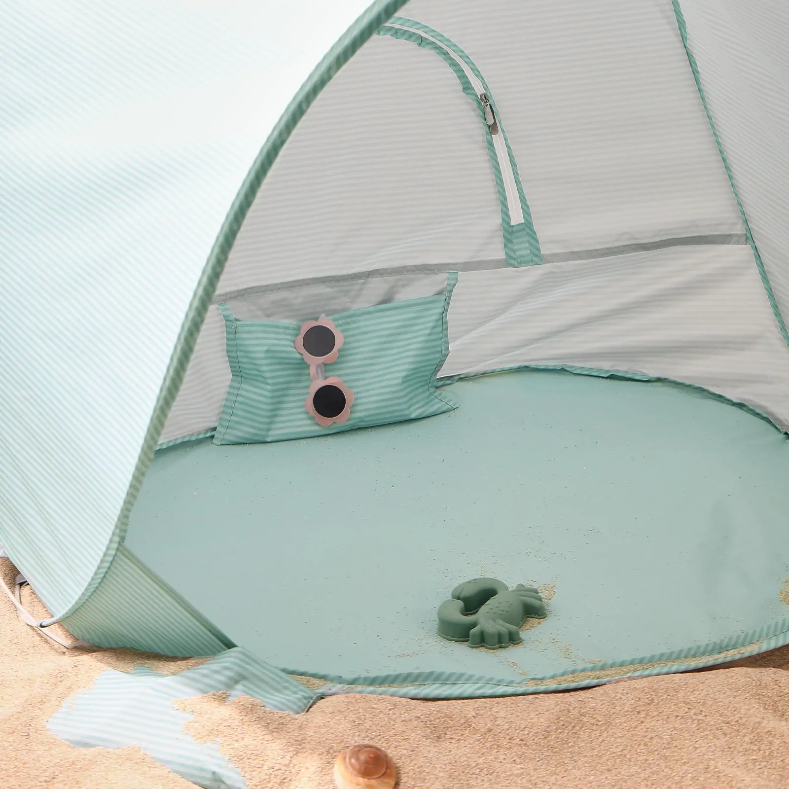 The Tiny Land® One-Stop Beach Toy & Tent Set – Sea Blue sits on the sand, complete with a matching pillow, round pink sunglasses, and a green toy turtle from Tiny Land. Some sand is scattered inside the UV-protective shade tent.