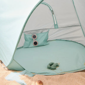The Tiny Land® One-Stop Beach Toy & Tent Set – Sea Blue sits on the sand, complete with a matching pillow, round pink sunglasses, and a green toy turtle from Tiny Land. Some sand is scattered inside the UV-protective shade tent.