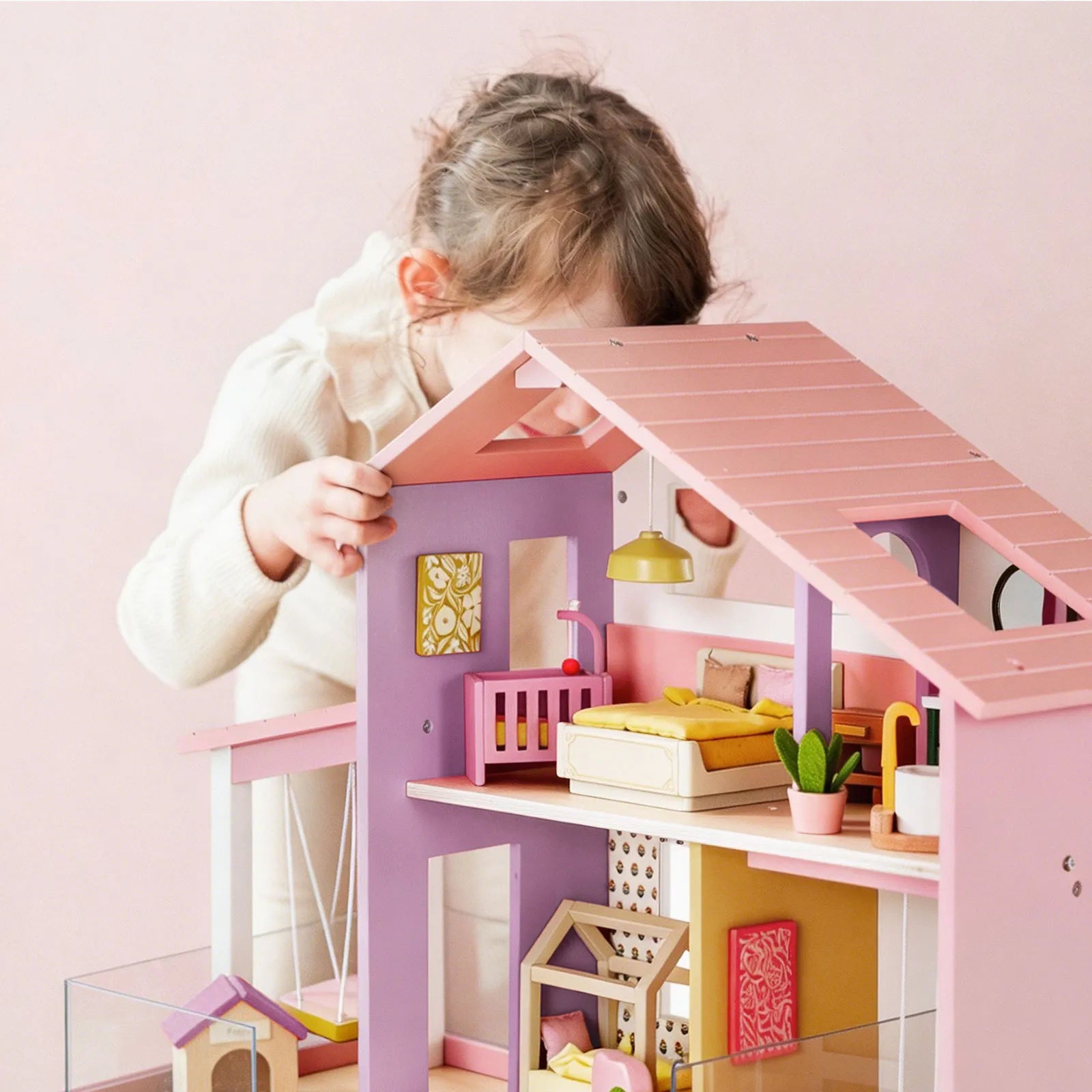 A young child in a white top plays with the Tiny Land® Sweetwood Cottage Dollhouse, featuring pink and purple walls, miniature furniture, and decor, set against a pale pink background.|Pink