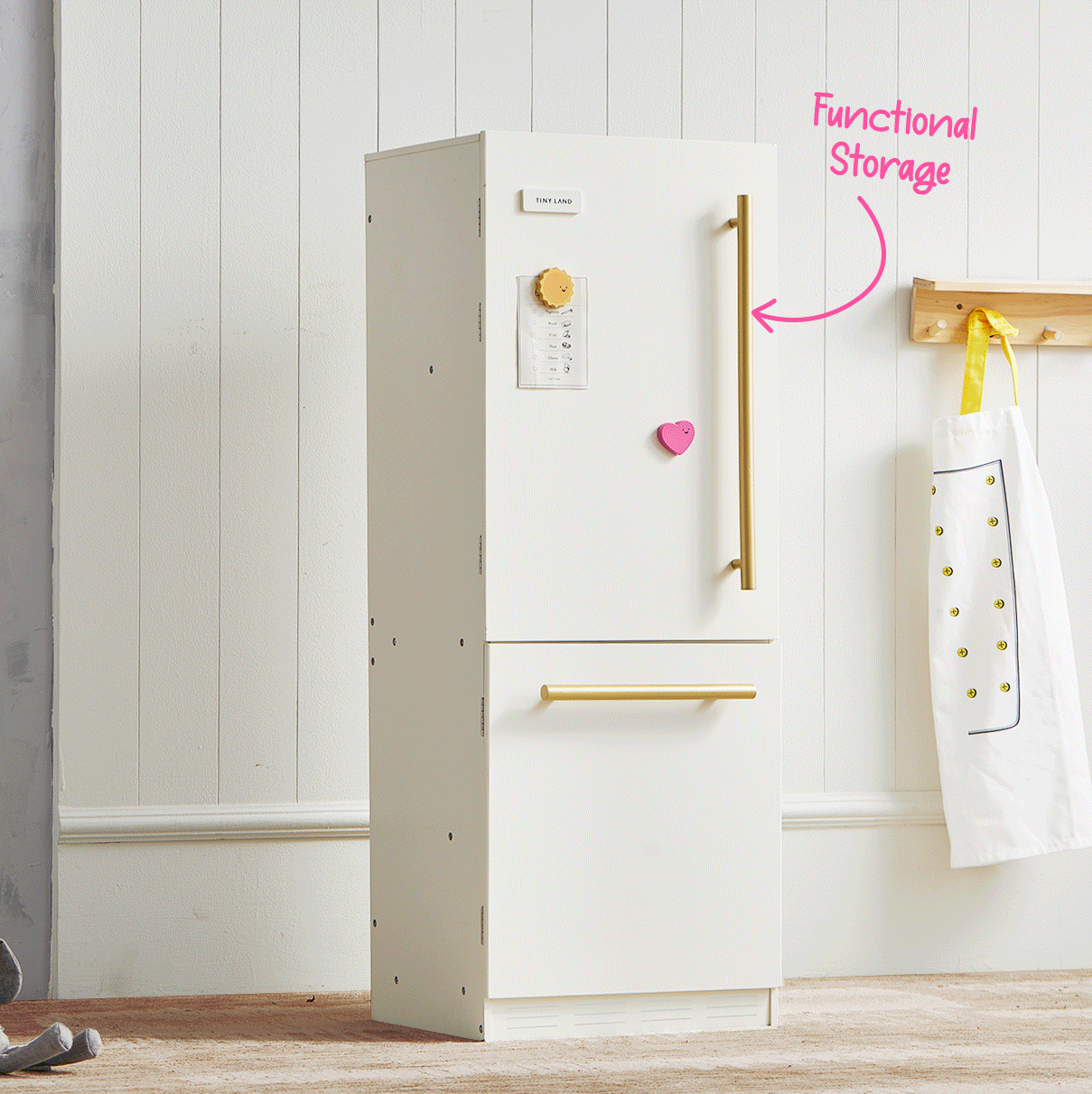 A white Tiny Land® BistroSwitch Kitchen & Fridge Set with gold handles stands against a white wall. An apron hangs nearby, and pink text with an arrow points to the fridge, reading Functional Storage—making this Tiny Land set a stylish addition to any kid’s play kitchen or pretend play area.