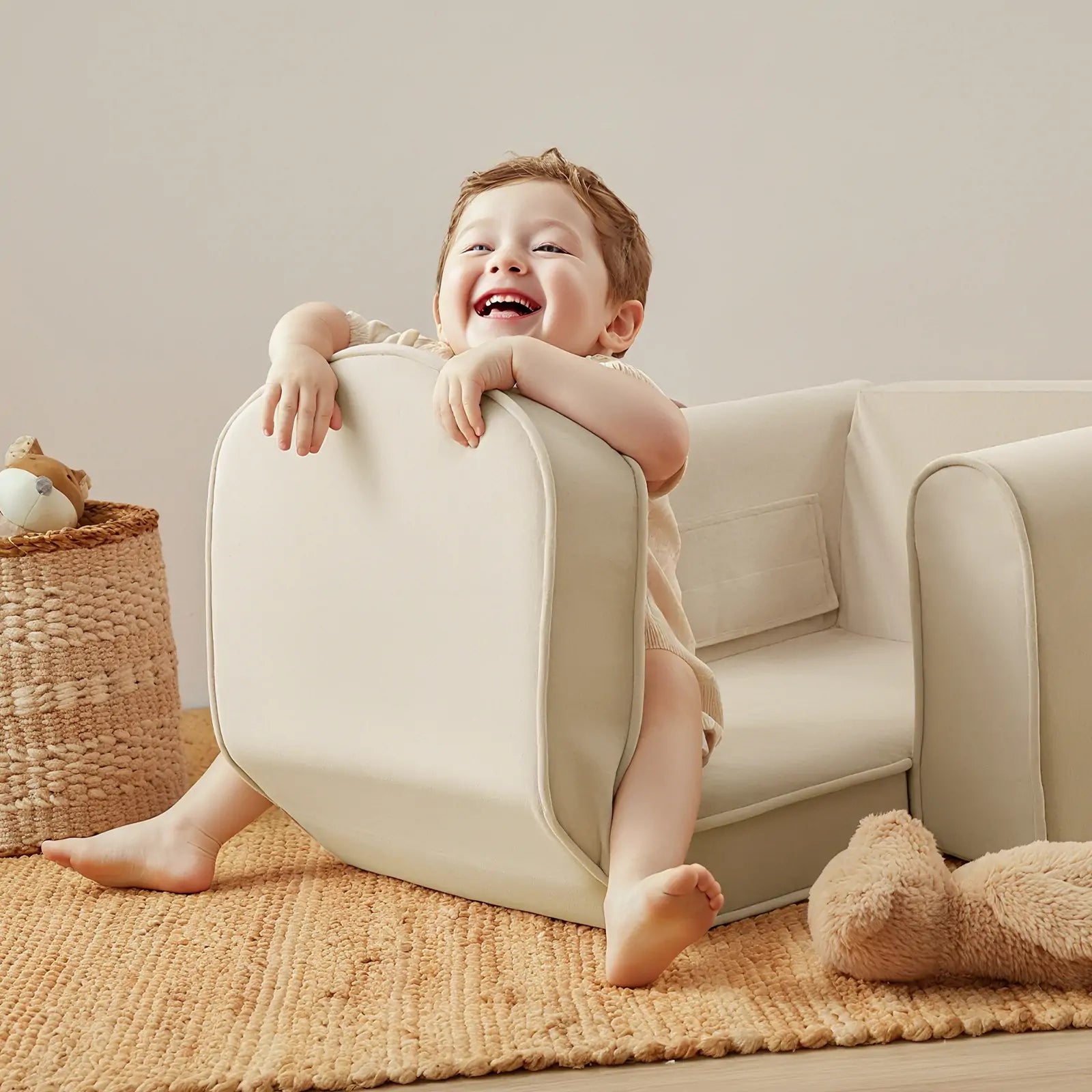 A smiling toddler sits playfully on a light beige couch in a cozy room, hugging the cushion of their Tiny Land® GentleSnug Kid Chair. A woven basket with toys and a large plush bear are nearby on a tan rug.|Beige
