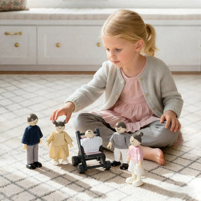 A young girl in a pink dress and gray cardigan sits on a carpet, playing with Tiny Land® Wooden Dollhouse Dolls by Tiny Land INC—five poseable wooden dolls, one in a stroller—in a bright, cozy room.