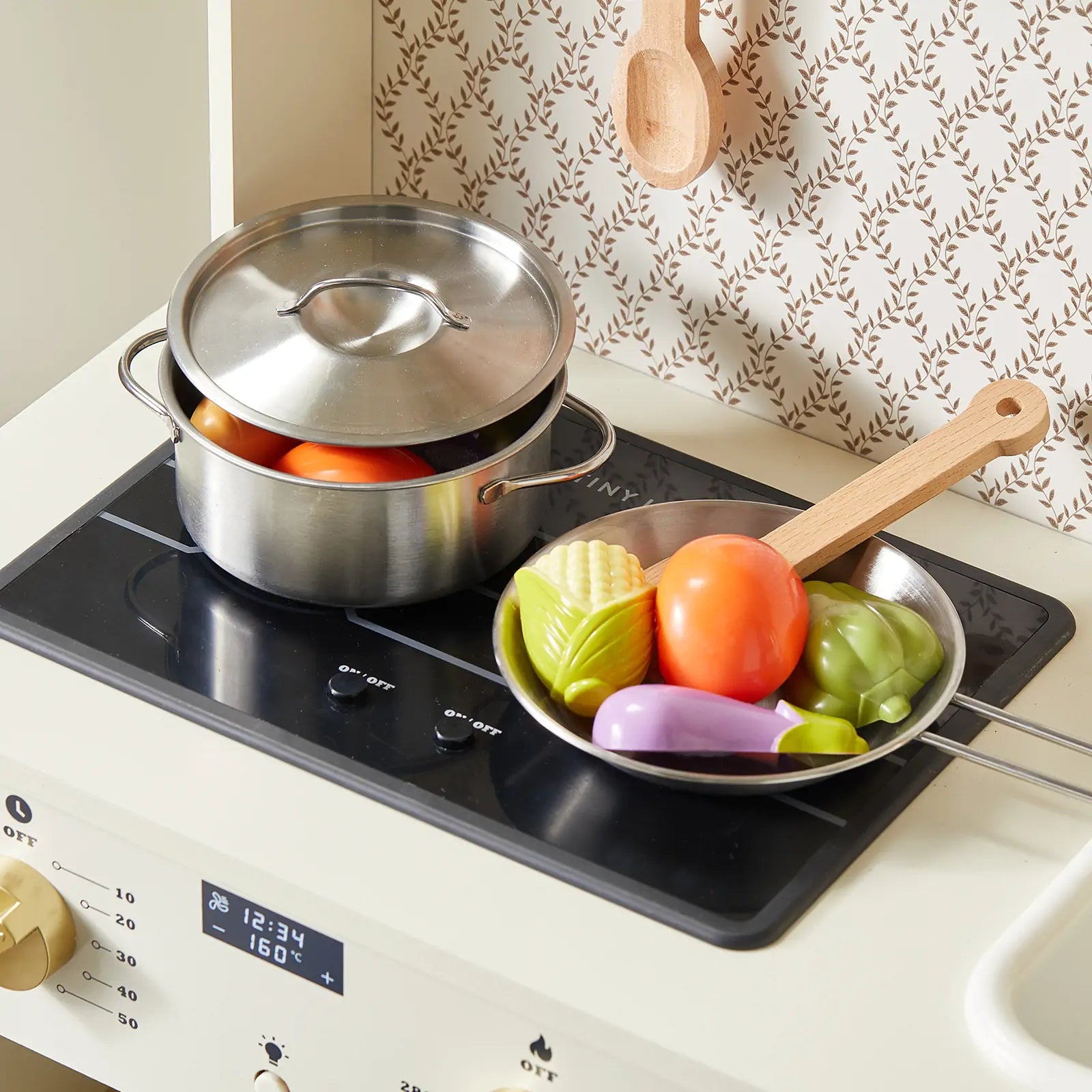 The Tiny Land® PopJoy Play Kitchen by Tiny Land features a wooden stove, metal pot with toy vegetables, a frying pan, wooden spatula, patterned wallpaper, and hanging spoon—perfect for creative pretend play.