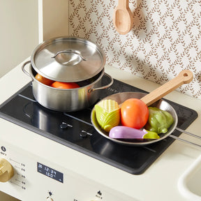 The Tiny Land® PopJoy Play Kitchen by Tiny Land features a wooden stove, metal pot with toy vegetables, a frying pan, wooden spatula, patterned wallpaper, and hanging spoon—perfect for creative pretend play.