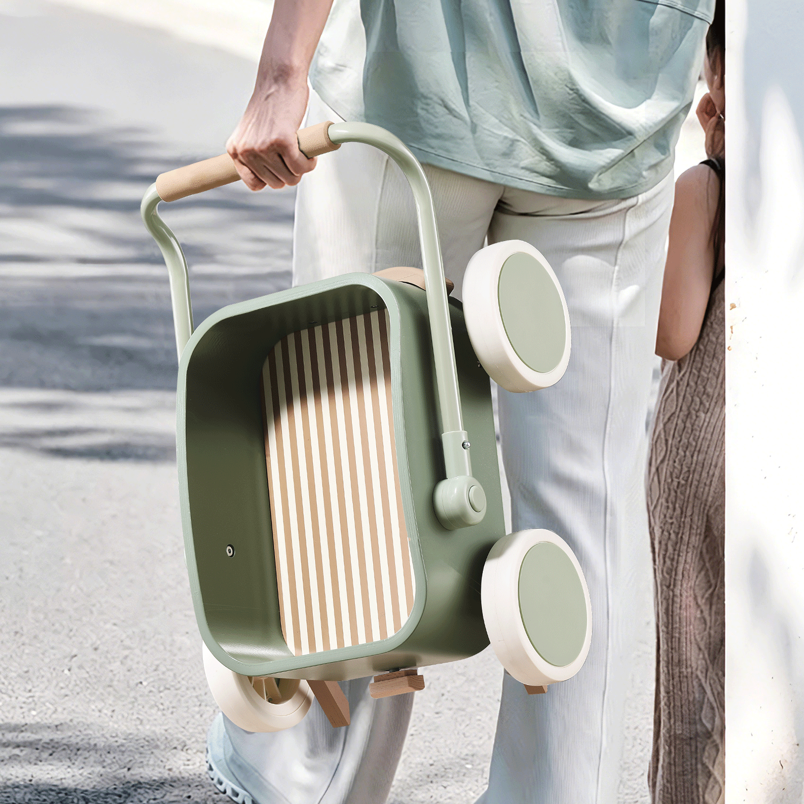 A person holds the Tiny Land® JoyRide Baby Walker with Blocks - Sage by Test, walking outdoors with a child whose face is partially hidden behind a wall on a sunny day.