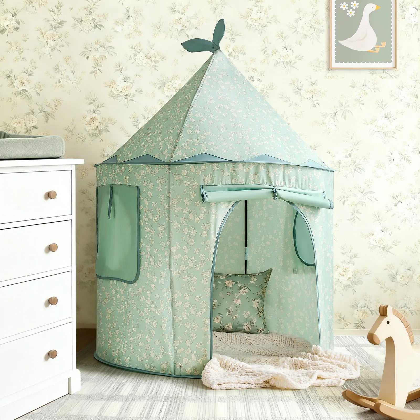 The Tiny Land® Tiny Sprout Castle Tent in mint green, featuring a pointed roof and leaf accents, stands in a child’s room with floral wallpaper, near a wooden rocking horse, white dresser, and framed goose picture on the wall.