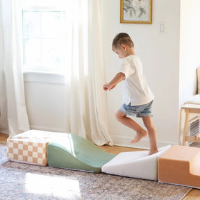 A young boy in a white shirt and denim shorts jumps barefoot on Tiny Land® PlayNest Climbing Blocks by Tiny Land INC, arranged like stepping stones in a bright, sunlit room with white curtains and a rug, encouraging gross motor development.