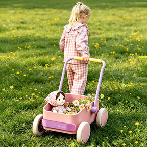 A young child in pink checkered pajamas pushes the Tiny Land® 3-in-1 Wooden Baby Walker Wagon - Pink by Tiny Land, carrying a doll and wildflowers, across a grassy field scattered with yellow and white flowers.