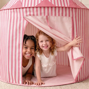 Two young girls smile as they play inside the Tiny Land® Kids Tent – Indoor Tent for Toddlers by Tiny Land. One girl holds open the pink and white striped flap while both peek out, looking happy and playful. The kids tent is set up on a light-colored rug indoors.|Pink
