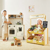 Two kids in chef hats enjoy their Tiny Land® Farm-to-Table Gift Set, featuring a light & sound kitchen and market stall, play food, and market signs for imaginative cooking and selling fun.