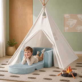 A young child smiles while relaxing in the Tiny Land® GentleSnug Corduroy Kid Chair by Tiny Land—a plush blue toddler chair nestled inside a white play tent. Nearby sits a toy campfire, and the room is styled in soothing neutral tones with nature-themed wall art and a dotted rug.|Soft Teal