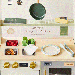 The Tiny Land® BistroSwitch Play Kitchen by Tiny Land includes a pan, utensils, faux veggies, plate, recipe cards, stovetop with dials and timer, and an accessory shelf. Great for pretend play! The text reads “100% Organic Tiny Kitchen.”.|Green