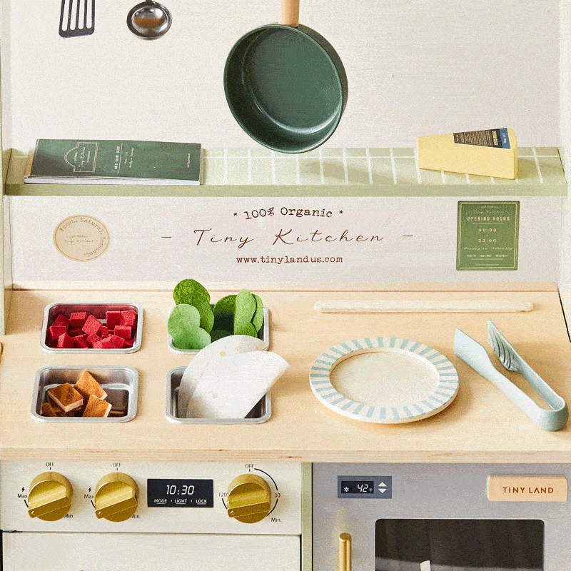 The Tiny Land® BistroSwitch Play Kitchen by Tiny Land includes a pan, utensils, faux veggies, plate, recipe cards, stovetop with dials and timer, and an accessory shelf. Great for pretend play! The text reads “100% Organic Tiny Kitchen.”.