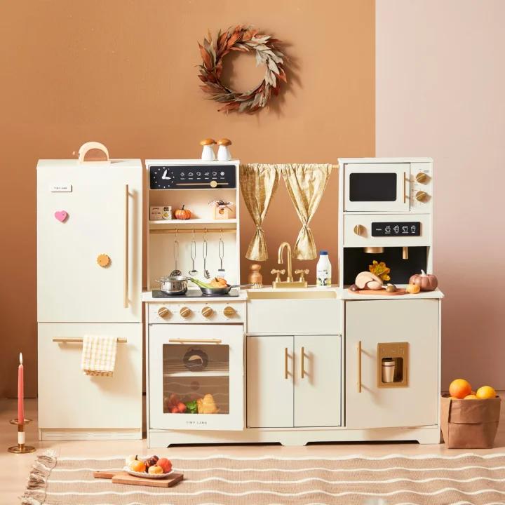 The Tiny Land All-in-One Family Feast Gift Set features a cream wooden play kitchen with fridge, oven, sink, microwave, and stove, plus toy food and utensils. Set against a pink-beige wall with a wreath and candle. Ideal for pretend play!.