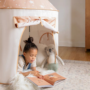 A young child lies on a soft rug inside the star-patterned Tiny Land® Adventure 3-in-1 Play Tunnel & Ocean Ball Set by Tiny Land, reading a picture book with a stuffed bear toy beside them. This cozy play set includes a tent, an exciting play tunnel, and a ball pit with ocean balls for hours of imaginative fun. The wooden floors and plush rug create the perfect space for endless Tiny Land adventures.