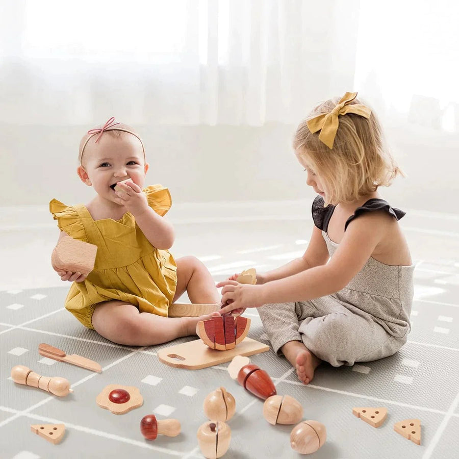 Two children play on a mat with the Tiny Land® Play Kitchen Accessories – Play Food for Play Kitchen: Wooden Cut and Play Food Toys by Tiny Land Play Food, smiling and focused as sunlight shines through sheer curtains.