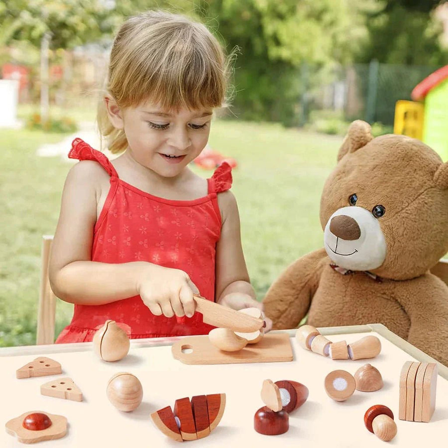 A young child in a red dress plays outdoors with the Tiny Land Play Food Wooden Cut and Play Food Toys set and a pretend knife at a table, alongside a large brown teddy bear. Trees and playground equipment can be seen in the background.