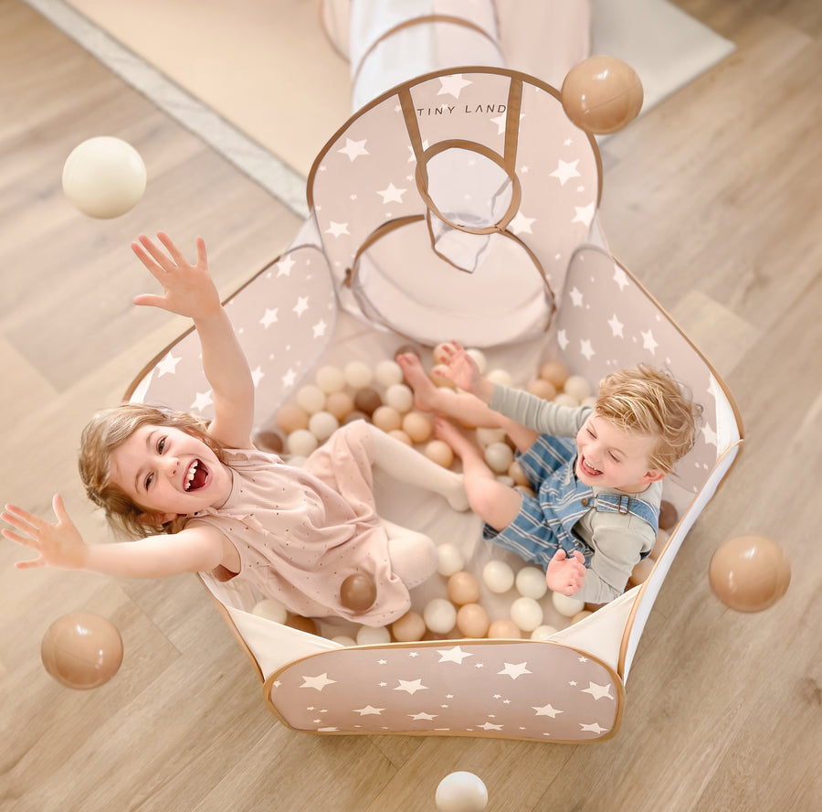 Two young children laugh and toss Tiny Land® Ocean Ball - 200 pcs with Varied Colors in a star-patterned, beige playpen filled with soft, non-toxic balls. A play tunnel attaches to the playpen on a light wood floor. Brand: Tiny Land.|Brown