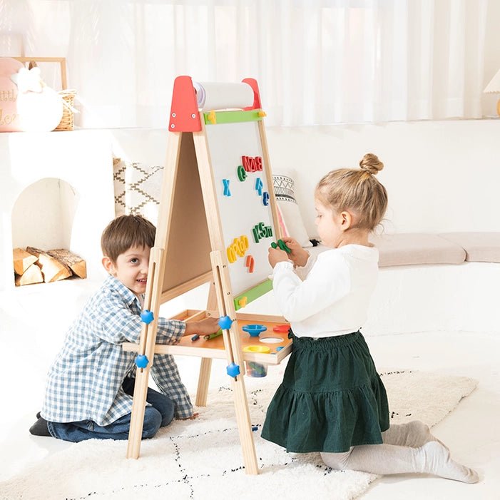 A young boy and girl play in a cozy room with a Tiny Land® Easel and arrange colorful magnets on the board, while the boy smiles over a roll of Tiny Land® Easel Paper Roll (3 rolls).