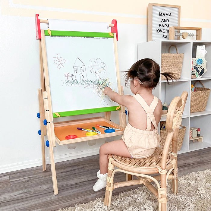 A small child draws with markers and crayons on Tiny Land® Easel Paper Roll (3 rolls) at a double-sided easel, sketches of a house, tree, and sun displayed. Shelves with baskets and toys appear in the background.