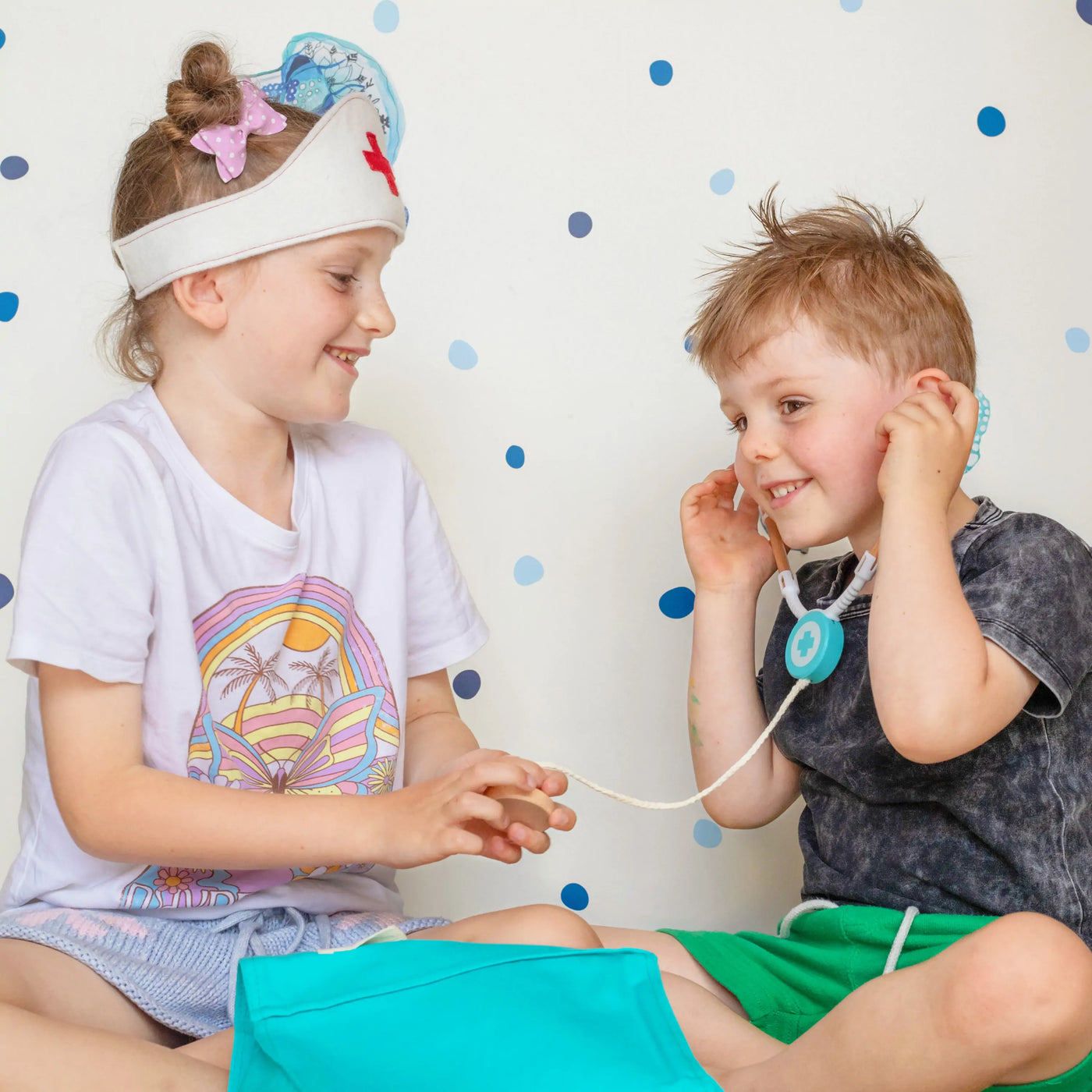Two smiling children play together on the floor as the girl, wearing a nurse headband, pretends to check the boy using the Tiny Land® Doctor Kit for Kids by Tiny Land. The boy holds a toy stethoscope against a dotted background.