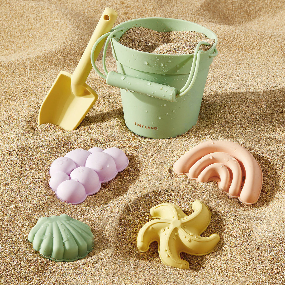 The Tiny Land® Sand-Buddies Sand Toy Set by Tiny Land INC features a pastel green bucket, yellow shovel, and four silicone beach toys—a seashell, starfish, banana, and grapes—arranged on sand for engaging sensory play.