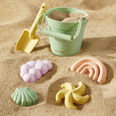 The Tiny Land® Sand-Buddies Sand Toy Set by Tiny Land, featuring a green bucket, yellow plastic shovel, and four pastel sand molds, sits on the sand—perfect for sensory and creative play at the beach.