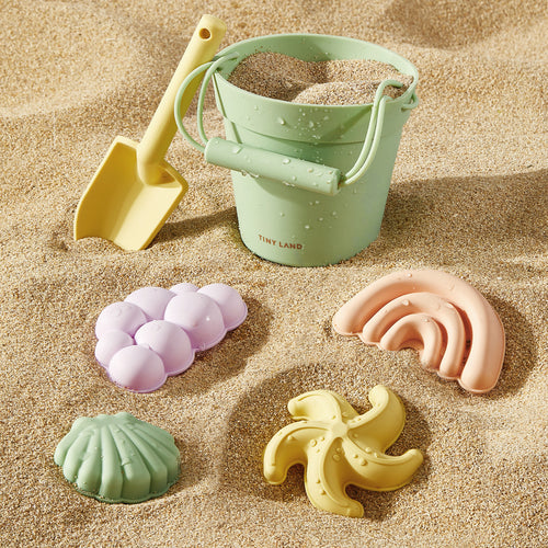 The Tiny Land® Sand-Buddies Sand Toy Set by Tiny Land INC features a pastel green bucket, yellow shovel, and four silicone beach toys—a seashell, starfish, banana, and grapes—arranged on sand for engaging sensory play.