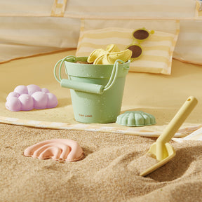 A pastel green bucket from the Tiny Land® Sand-Buddies Sand Toy Set sits on the beach with pastel rainbow and cloud molds, plus a yellow spade. A striped pillow and sunglasses are visible in the background.