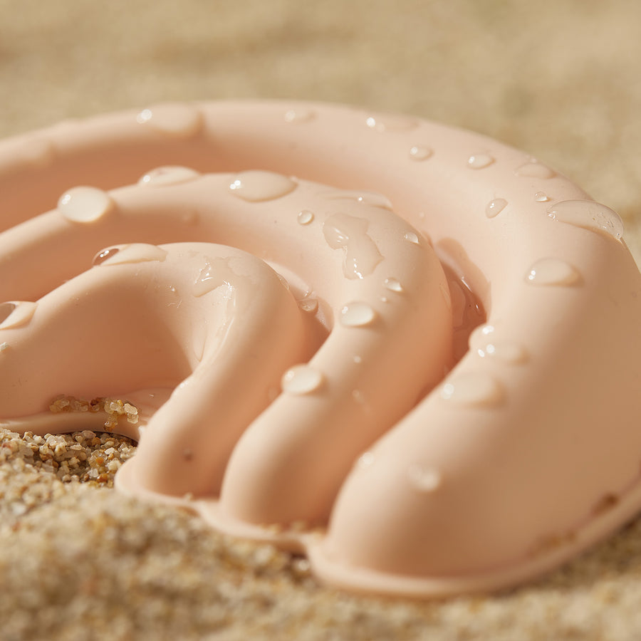 Close-up of a pale pink, wavy silicone toy from the Tiny Land® Sand-Buddies Sand Toy Set by Tiny Land, with water droplets and sand on its surface, lying on a sandy background.