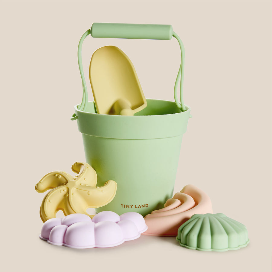 The Tiny Land® Sand-Buddies Sand Toy Set by Tiny Land features a green bucket with a yellow spade and pastel silicone toys shaped like a starfish, seashell, flower, and swirl—ideal for toddler sand play.
