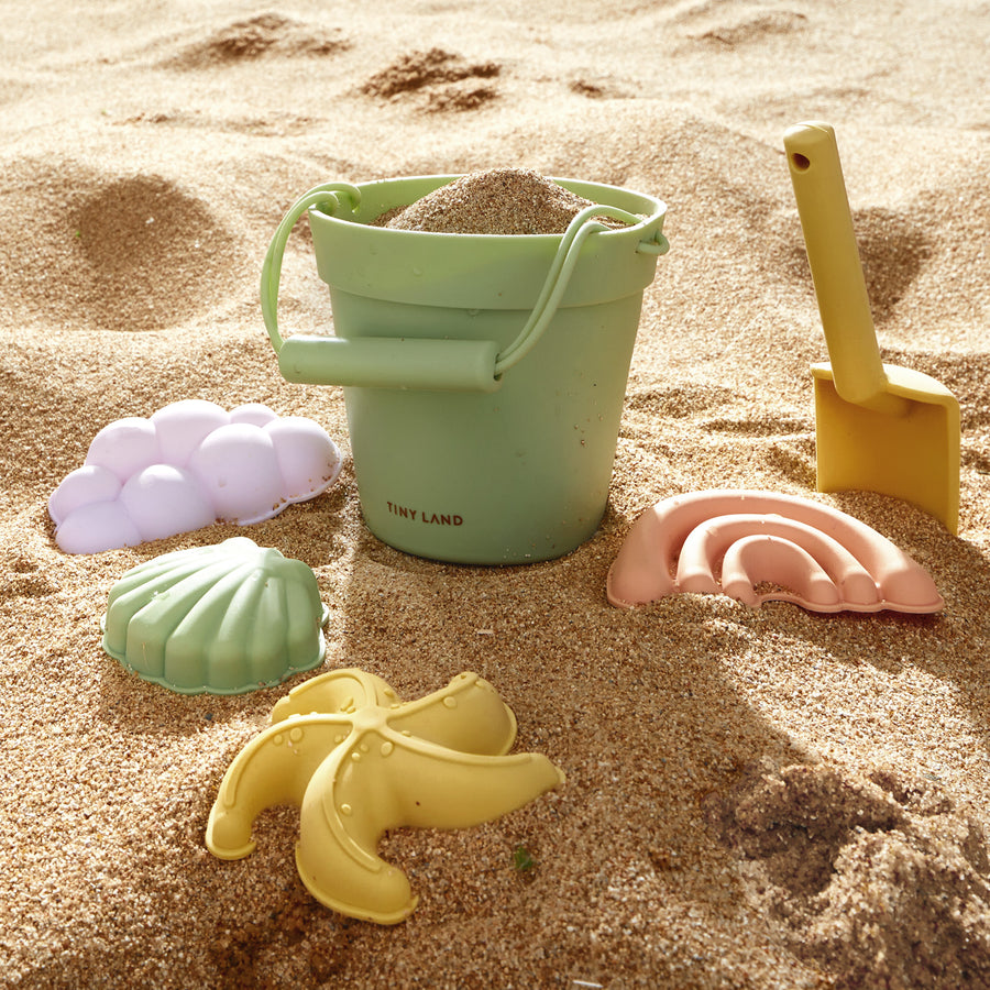 The Tiny Land® Sand-Buddies Sand Toy Set includes a green bucket with sand, a yellow shovel, and four pastel silicone molds—banana, seashell, shell, and rainbow—for endless toddler fun at the beach.