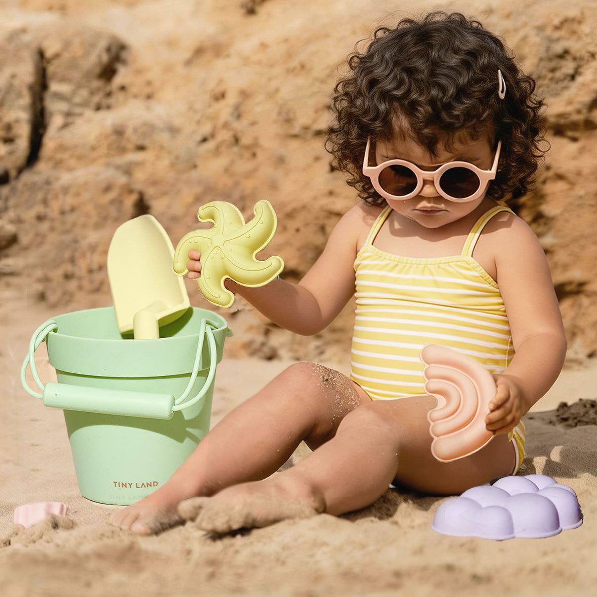 A young child in a yellow striped swimsuit and pink sunglasses plays with the Tiny Land® Sand-Buddies Sand Toy Set by Tiny Land INC on a sandy beach near a green bucket, with rocky terrain in the background.