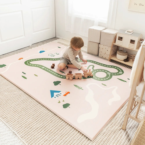 A young child sits on the Tiny Land® Double-Sided Baby Playmat Forest Track Wonder, a pink, waterproof GFOAM mat, playing with a wooden train in a bright, cozy room with pastel decor and neat storage cubes.