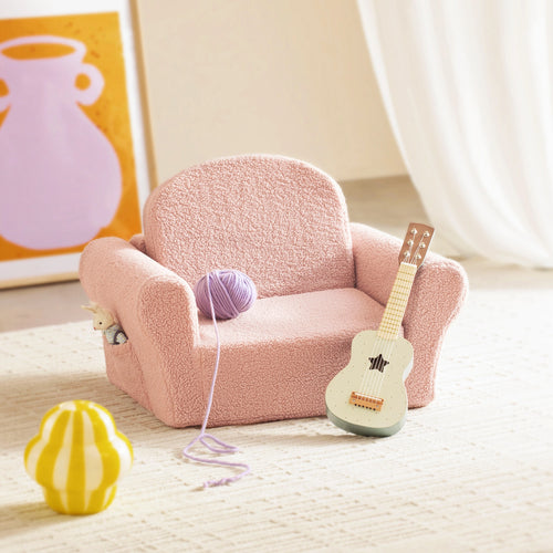 The Tiny Land® Afternoon Couch - Pink Kids Chair by Tiny Land sits on a light rug with a purple yarn ball and toy guitar. A striped yellow plush toy rests on the floor, while abstract art decorates the background wall.