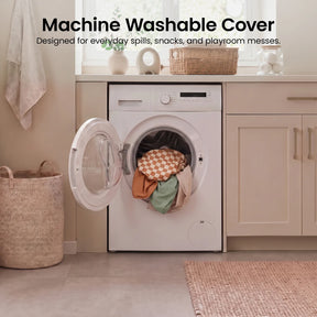 A washing machine with its door open, filled with colorful fabric—including pieces from the Tiny Land® PlayNest Climbing Blocks by Tiny Land. The room features beige cabinets, a woven basket, and a towel hanging on the wall. Text above reads Machine Washable Cover.