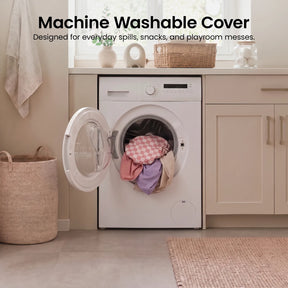 A front-load washing machine with colorful laundry inside sits in a bright, modern laundry room featuring neutral cabinets, a woven basket, and towels hanging nearby. In the corner, the Tiny Land® PlayNest Climbing Blocks - Violet by Tiny Land provide a fun play space for little ones to enjoy after playtime. Text above reads: Machine Washable Cover.