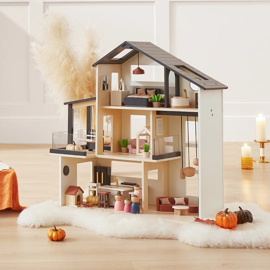The Tiny Land® Sweetwood Edge Dollhouse by Tiny Land is displayed on a fluffy white rug with small pumpkins, candles, and pampas grass for a cozy autumn vibe. Miniature family models complete this inviting scene.