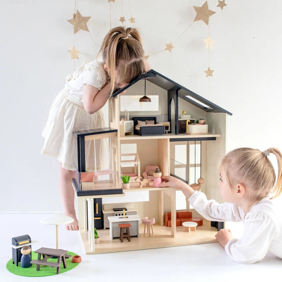 Two young girls in white dresses play with the Tiny Land® Sweetwood Edge Dollhouse. One stands peering in from above, while the other sits reaching into its miniature rooms. Star decorations hang in the background.