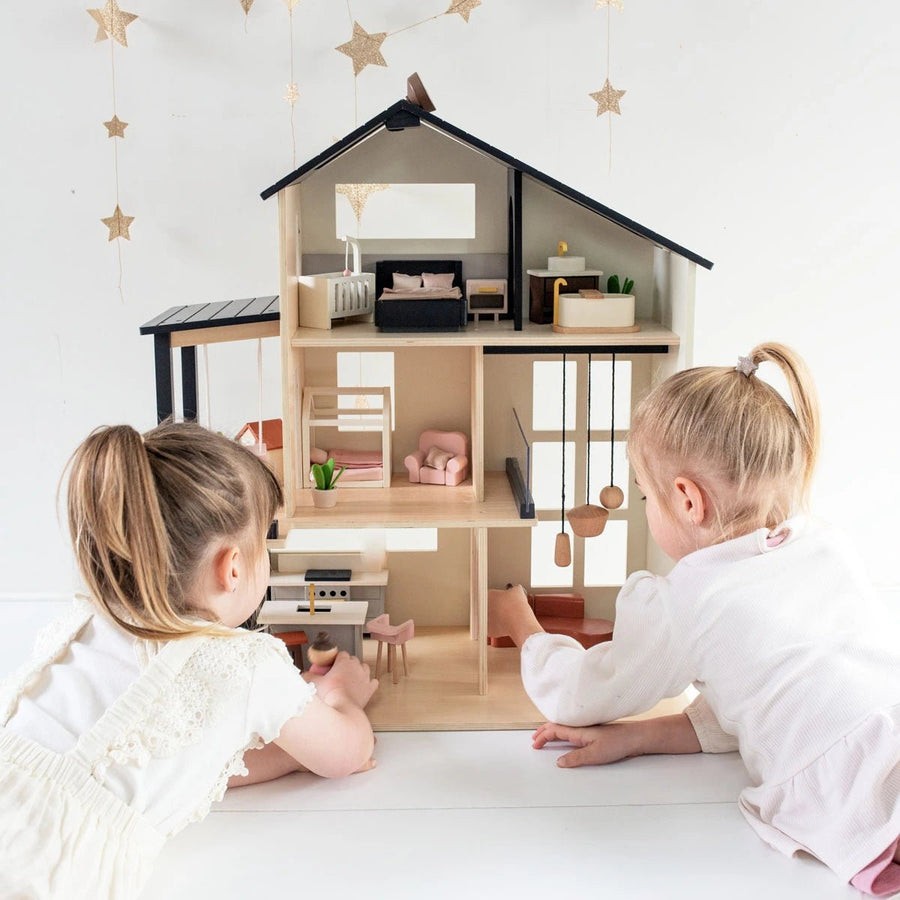 Two young girls with light hair play on the floor with the Tiny Land® Sweetwood Edge Dollhouse, featuring miniature furniture and family figures, beneath star-shaped decor on a white wall. Brand: Tiny Land.