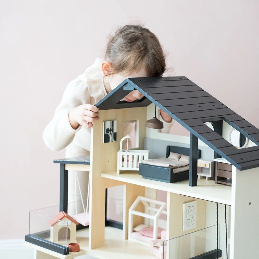 A young child with light brown hair, in a light sweater, leans over to look inside the Tiny Land® Sweetwood Edge Dollhouse by Tiny Land, featuring miniature furniture and family models against a pale pink background.