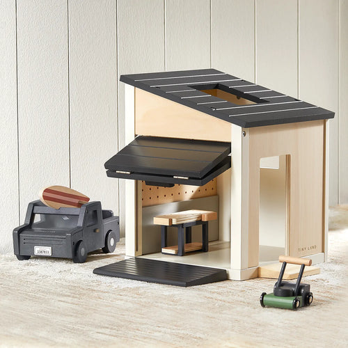 A Tiny Land® Mystery Surprise Box showcases a wooden toy garage with a black roof and open door, paired with premium toys—a black truck, wooden car, and green lawnmower—on a light surface against white paneling.