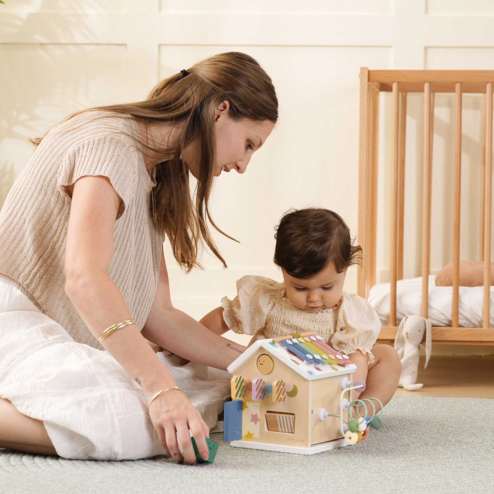 A woman and a child play with the Tiny Land® Cottage Activity Cube - Morandi by Tiny Land in a softly lit nursery, with a crib visible in the background.